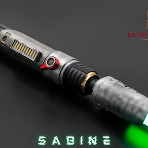 May include: A metallic lightsaber with a green blade, featuring a silver and black handle with gold and red accents. The word "SABINE" is displayed in green digital font. The Astro MX Sabers logo is in the upper right corner.