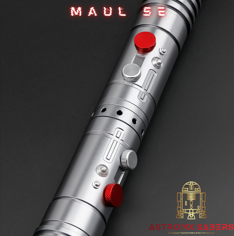 Op de afbeelding: Een zilveren lichtzwaard met rode accenten, met de tekst "MAUL SE" in het rood. Het lichtzwaard heeft een metallic afwerking met diverse knoppen en details. Het AstroX Sabers-logo is rechtsonder zichtbaar.