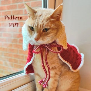 May include: A ginger cat wearing a red and white crocheted cape with a white trim and a red and white bow. The cat is sitting on a windowsill looking out the window. The text "Pattern PDF" is visible in the top left corner of the image.