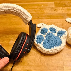 May include: A black headset with a white crocheted headband. Two crocheted paw print designs, one blue and one orange, are in the background.