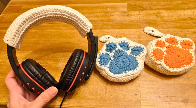 May include: A black headset with a white crocheted headband. Two crocheted paw print designs, one blue and one orange, are in the background.