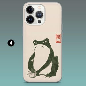 Pode incluir: Capa de telem&oacute;vel com um design de inspira&ccedil;&atilde;o japonesa. Apresenta um sapo verde escuro com uma express&atilde;o zangada, segurando uma tigela. O design inclui um selo vermelho e uma est&eacute;tica minimalista. A capa tem uma borda transparente e um fundo bege.