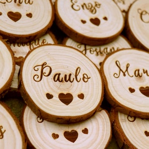 Peut inclure: Tranches de bois avec des noms gravés en écriture cursive et des motifs de cœur. Les noms incluent "Paulo", "Marie", "Enzo" et "Océan". Le bois est de couleur brun clair naturel. Parfait pour les mariages.