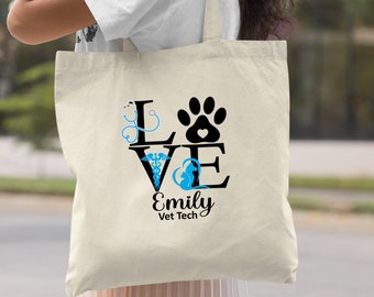 Puede incluir: Una bolsa de lona natural con un diseño en negro y azul que dice "LOVE" con una huella de pata y un estetoscopio. El texto "Emily Vet Tech" está debajo del diseño.