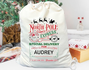May include: A white canvas Santa sack with a red and green North Pole Express design. The sack has a drawstring closure and says "Special Delivery for Audrey" and "Do Not Open Until December 25th".
