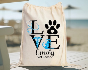 May include: A white canvas drawstring bag with the word "LOVE" in black letters. The "L" is decorated with a blue stethoscope, the "V" with a caduceus, the "O" with a paw print, and the "E" with a silhouette of two dogs. The text "Emily Vet Tech" is printed below the word "LOVE".