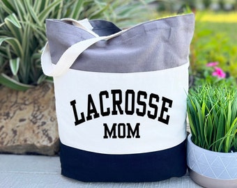 May include: A tri-color grey canvas tote bag with black handles and the text "Lacrosse Mom" printed in black on a white stripe.