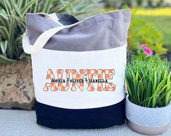 May include: A grey and white tote bag with a black bottom. The bag has a checkered design with the word "AUNTIE" in orange letters. The names "Sophia", "Oliver", and "Isabella" are written in smaller letters below the word "AUNTIE".