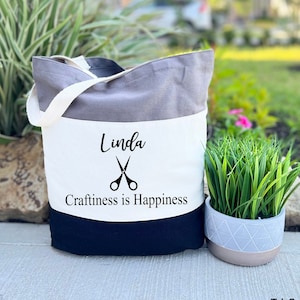 May include: A tri-color tote bag with a gray, white, and black design. The bag has a white panel with the name "Linda" and the text "Craftiness is Happiness" with a pair of scissors graphic.