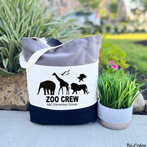 May include: A tri-color tote bag with a white panel featuring a black silhouette of a giraffe, elephant, lion, and two birds. The text "Zoo Crew" and "ABC Elementary School" is printed in black below the animals.