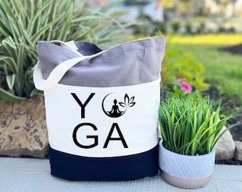 May include: A tri-color tote bag with a grey, white, and black design. The bag has the word "YOGA" printed on it in black with a silhouette of a person meditating in a lotus pose with a crescent moon and a flower.