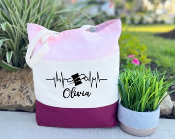 May include: A tri-color tote bag with a pink, white, and burgundy design. The bag features a black and white graphic of a sewing needle and thread with a heartbeat line and the name "Olivia" printed on the front.