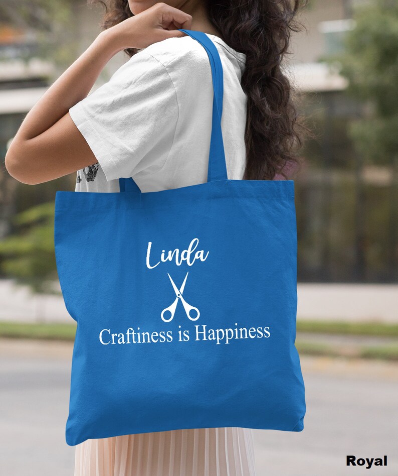 May include: A royal blue tote bag with white text that reads "Linda" with a pair of scissors and "Craftiness is Happiness".