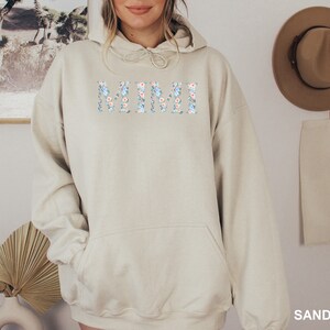 May include: A sand-colored hoodie with the word "MIMI" across the chest in a floral design of blue and peach flowers. The hoodie has a drawstring hood and a front pocket. The word "SAND" is printed at the bottom right corner.