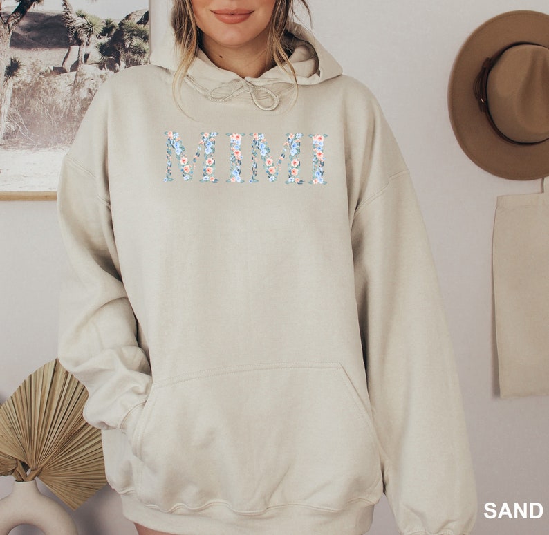 May include: A sand-colored hoodie with the word "MIMI" across the chest in a floral design of blue and peach flowers. The hoodie has a drawstring hood and a front pocket. The word "SAND" is printed at the bottom right corner.