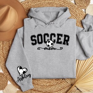 May include: Gray hooded sweatshirt with the text "Soccer Mom" and a soccer ball graphic. The left sleeve has a heart with a soccer ball and the name "Ashley" underneath.