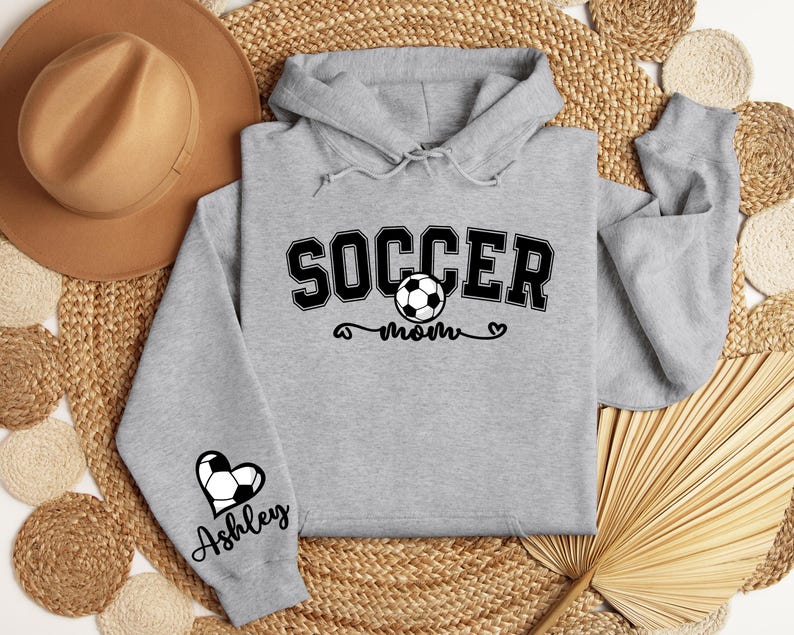 May include: Gray hooded sweatshirt with the text "Soccer Mom" and a soccer ball graphic. The left sleeve has a heart with a soccer ball and the name "Ashley" underneath.