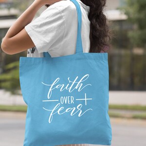 Może przedstawiać: Jasnoniebieska torba typu tote z napisem "faith over fear" w białym kolorze z symbolem krzyża.