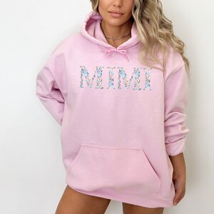 May include: A light pink hoodie with a front pocket and a drawstring hood. The word "MIMI" is displayed across the chest in a floral pattern with blue, white, and peach flowers. The hoodie is made of a soft material.