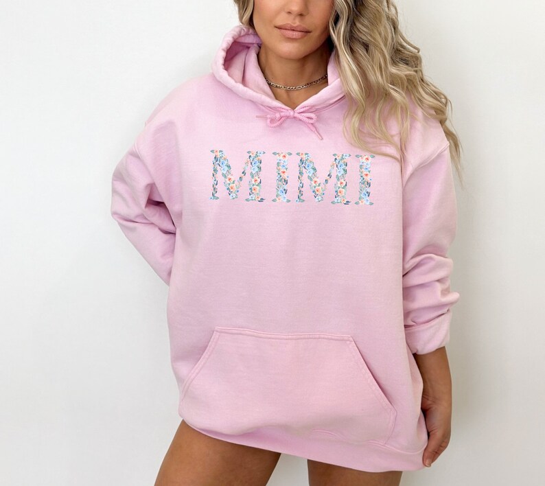 May include: A light pink hoodie with a front pocket and a drawstring hood. The word "MIMI" is displayed across the chest in a floral pattern with blue, white, and peach flowers. The hoodie is made of a soft material.