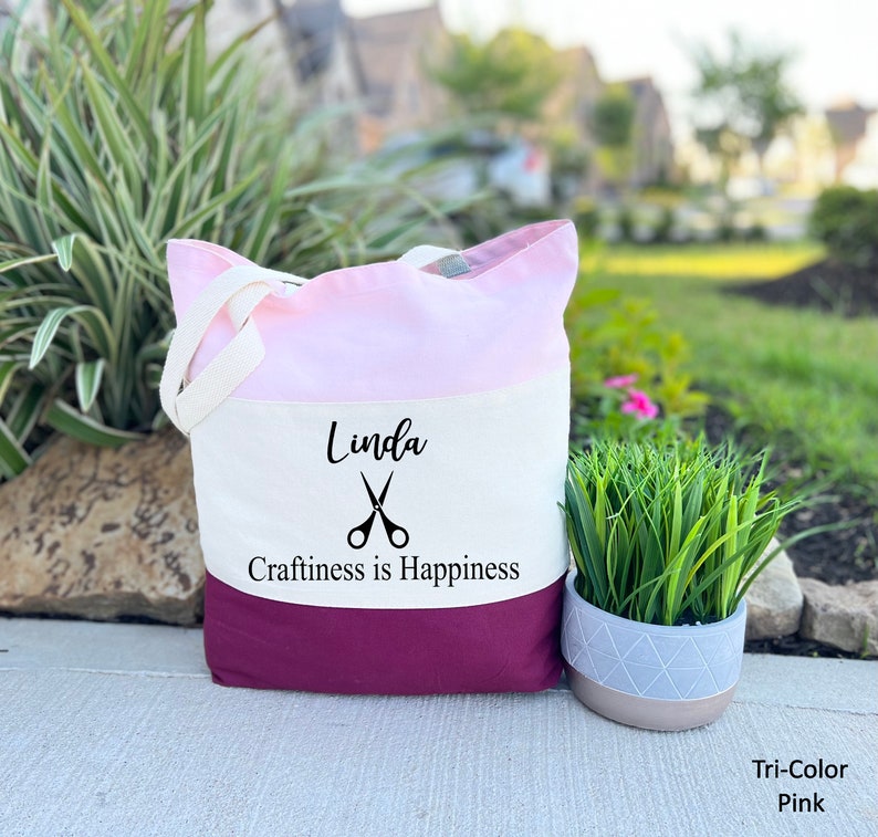 May include: A white canvas tote bag with a pink and burgundy ombre design. The bag has a black graphic of scissors and the text "Linda Craftiness is Happiness".