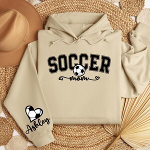 May include: Beige hoodie with a black graphic that says "Soccer Mom" with a soccer ball and a heart. The left sleeve has a black heart with a soccer ball and the name "Ashley" underneath.