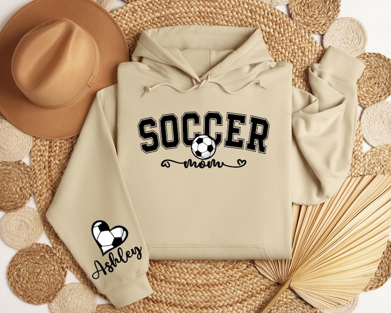May include: Beige hoodie with a black graphic that says "Soccer Mom" with a soccer ball and a heart. The left sleeve has a black heart with a soccer ball and the name "Ashley" underneath.