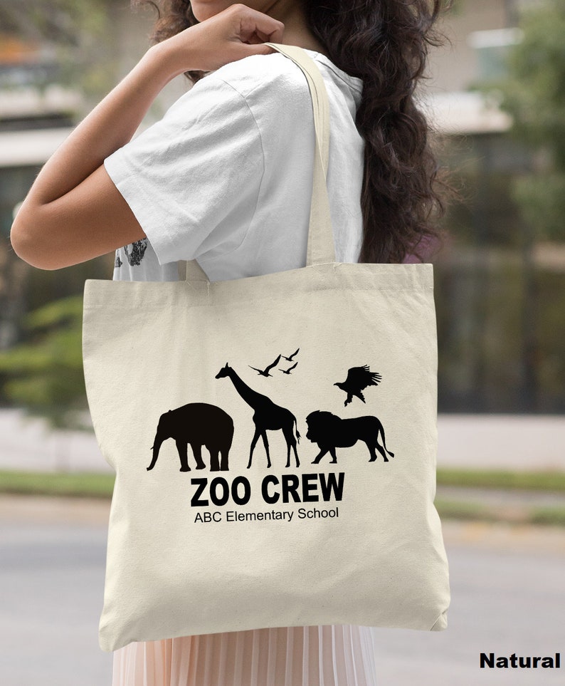 May include: A natural canvas tote bag with black silhouettes of animals, including an elephant, giraffe, lion, and birds. The text "Zoo Crew" and "ABC Elementary School" is printed in black.