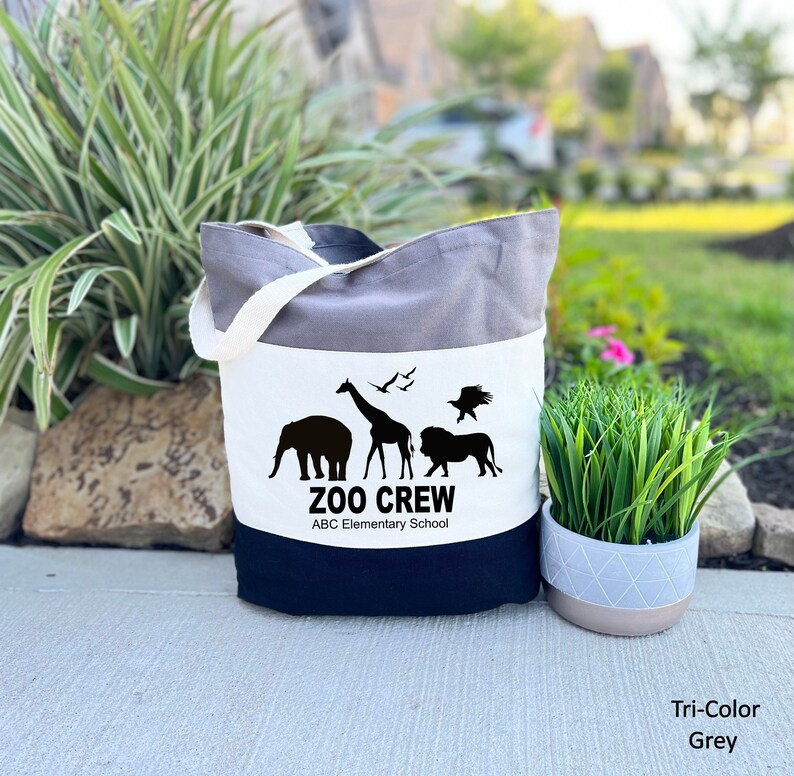May include: A tri-color tote bag with a white panel featuring a black silhouette of a giraffe, elephant, lion, and two birds. The text "Zoo Crew" and "ABC Elementary School" is printed in black below the animals.