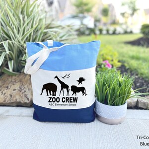 May include: A blue, white, and navy blue canvas tote bag with a white stripe and black animal silhouettes. The bag has the text "Zoo Crew" and "ABC Elementary School" printed on it.
