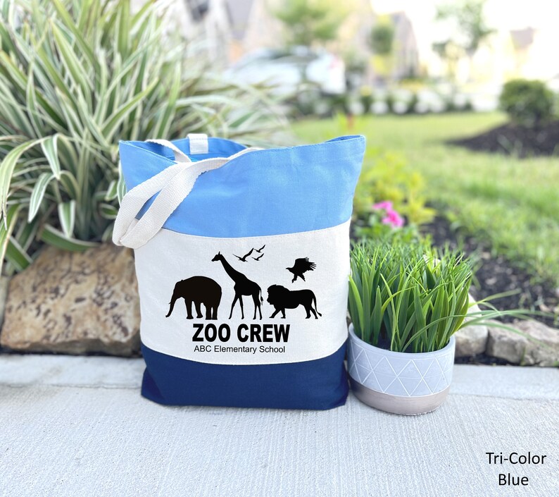 May include: A blue, white, and navy blue canvas tote bag with a white stripe and black animal silhouettes. The bag has the text "Zoo Crew" and "ABC Elementary School" printed on it.