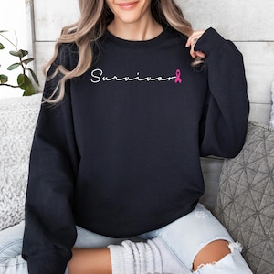 May include: Black sweatshirt with the word "Survivor" written in white script with a pink ribbon.