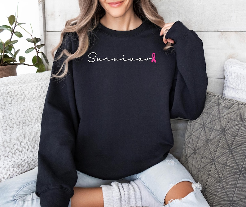 May include: Black sweatshirt with the word "Survivor" written in white script with a pink ribbon.