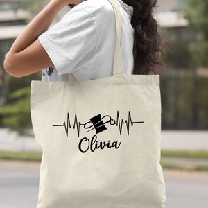 Puede incluir: Una bolsa de lona natural con un dise&ntilde;o negro de una aguja de coser y un carrete de hilo dentro de una l&iacute;nea de latido del coraz&oacute;n. El texto "Olivia" est&aacute; debajo del dise&ntilde;o.