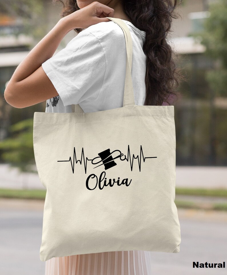 Puede incluir: Una bolsa de lona natural con un dise&ntilde;o negro de una aguja de coser y un carrete de hilo dentro de una l&iacute;nea de latido del coraz&oacute;n. El texto "Olivia" est&aacute; debajo del dise&ntilde;o.