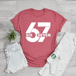 K&ouml;nnte beinhalten: Ein rotes T-Shirt mit der Zahl "67" in gro&szlig;er wei&szlig;er Schrift. Darunter steht "SIX SEVEN". Das Shirt wird mit Jeansshorts, einem wei&szlig;en Pullover und einer Kaffeetasse kombiniert.