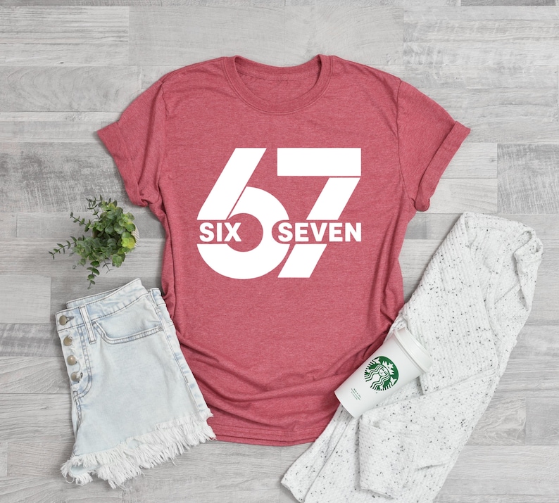 K&ouml;nnte beinhalten: Ein rotes T-Shirt mit der Zahl "67" in gro&szlig;er wei&szlig;er Schrift. Darunter steht "SIX SEVEN". Das Shirt wird mit Jeansshorts, einem wei&szlig;en Pullover und einer Kaffeetasse kombiniert.