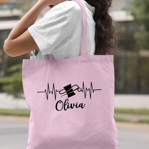 Puede incluir: Bolsa de tela de color rosa claro con un dise&ntilde;o de latido del coraz&oacute;n negro con tem&aacute;tica de costura y el nombre "Olivia" impreso en ella.