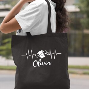 Puede incluir: Bolsa de tela negra con un dise&ntilde;o blanco de una aguja de coser y un hilo con una l&iacute;nea de latido del coraz&oacute;n y el nombre "Olivia" debajo.