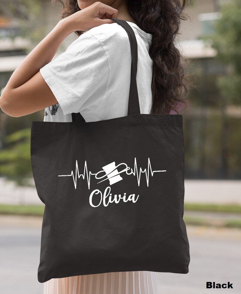 Puede incluir: Bolsa de tela negra con un dise&ntilde;o blanco de una aguja de coser y un hilo con una l&iacute;nea de latido del coraz&oacute;n y el nombre "Olivia" debajo.