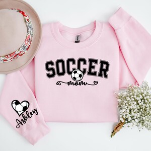 May include: Pink sweatshirt with the text "Soccer Mom" and a soccer ball graphic. The left sleeve has a heart with a soccer ball and the text "Ashley" on it.