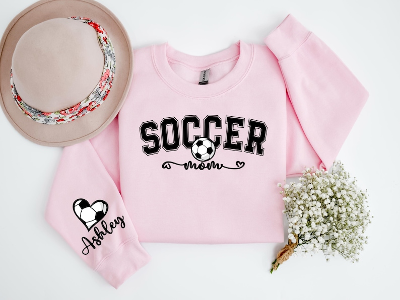 May include: Pink sweatshirt with the text "Soccer Mom" and a soccer ball graphic. The left sleeve has a heart with a soccer ball and the text "Ashley" on it.