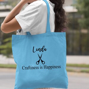 May include: Light blue tote bag with the name "Linda" and the text "Craftiness is Happiness" printed in black. A pair of scissors is printed above the text.