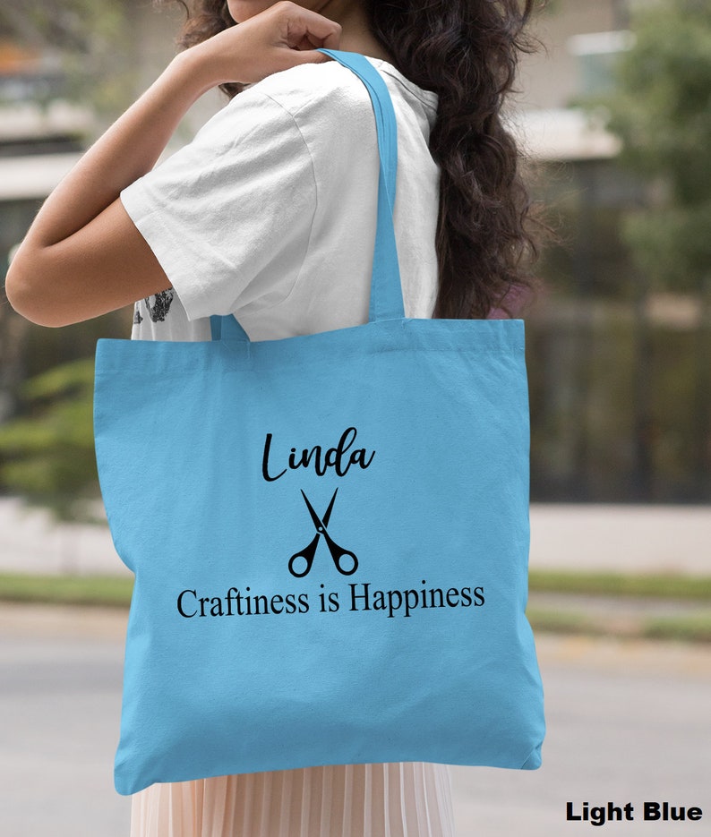 May include: Light blue tote bag with the name "Linda" and the text "Craftiness is Happiness" printed in black. A pair of scissors is printed above the text.