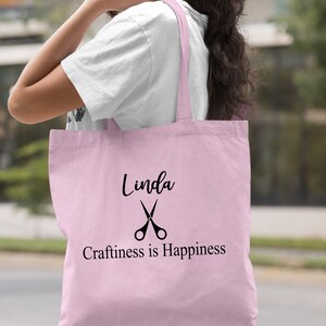 May include: Light pink tote bag with black text and a pair of scissors graphic. The text reads "Linda Craftiness is Happiness".