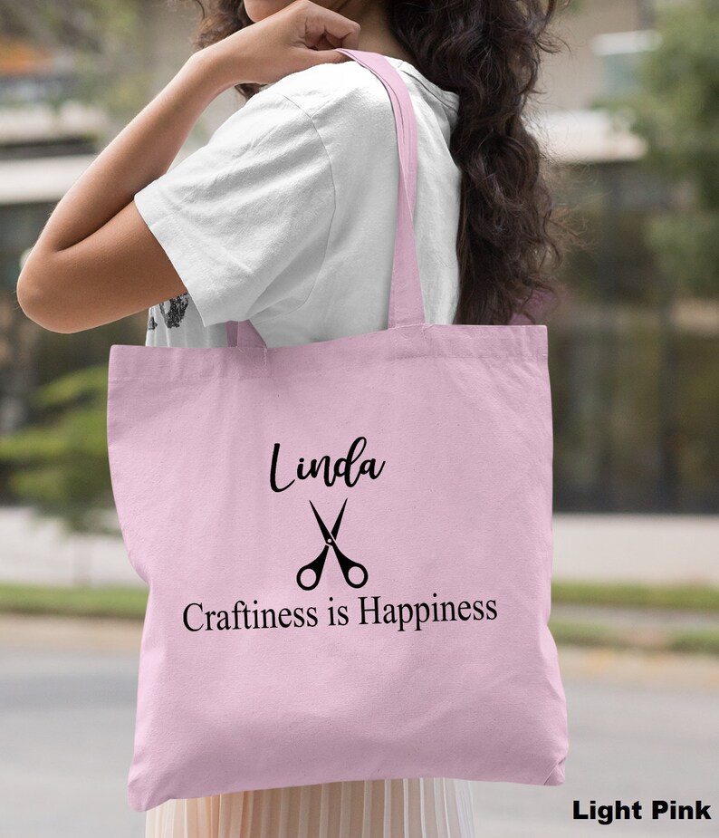 May include: Light pink tote bag with black text and a pair of scissors graphic. The text reads "Linda Craftiness is Happiness".