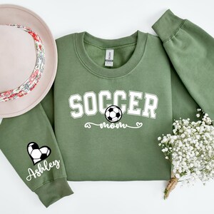 May include: Green sweatshirt with the text "Soccer Mom" and a soccer ball graphic. The left sleeve has a heart with a soccer ball and the text "Ashley".