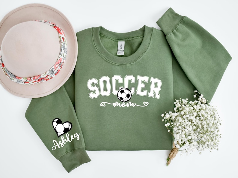May include: Green sweatshirt with the text "Soccer Mom" and a soccer ball graphic. The left sleeve has a heart with a soccer ball and the text "Ashley".