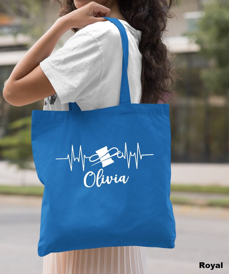 Puede incluir: Una bolsa de tela azul real con un dise&ntilde;o blanco de una aguja de coser y un hilo con una l&iacute;nea de latido del coraz&oacute;n y el nombre "Olivia".