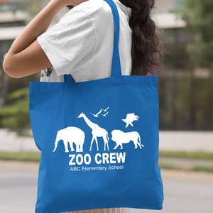 May include: A royal blue tote bag with white silhouettes of animals, including an elephant, giraffe, lion, and birds. The text "Zoo Crew" and "ABC Elementary School" is printed in white on the bag.